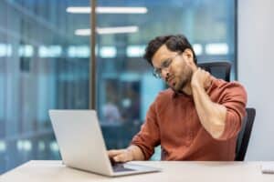 Man holding neck in pain while using laptop in office, highlighting workplace strain and chronic neck discomfort from prolonged computer use